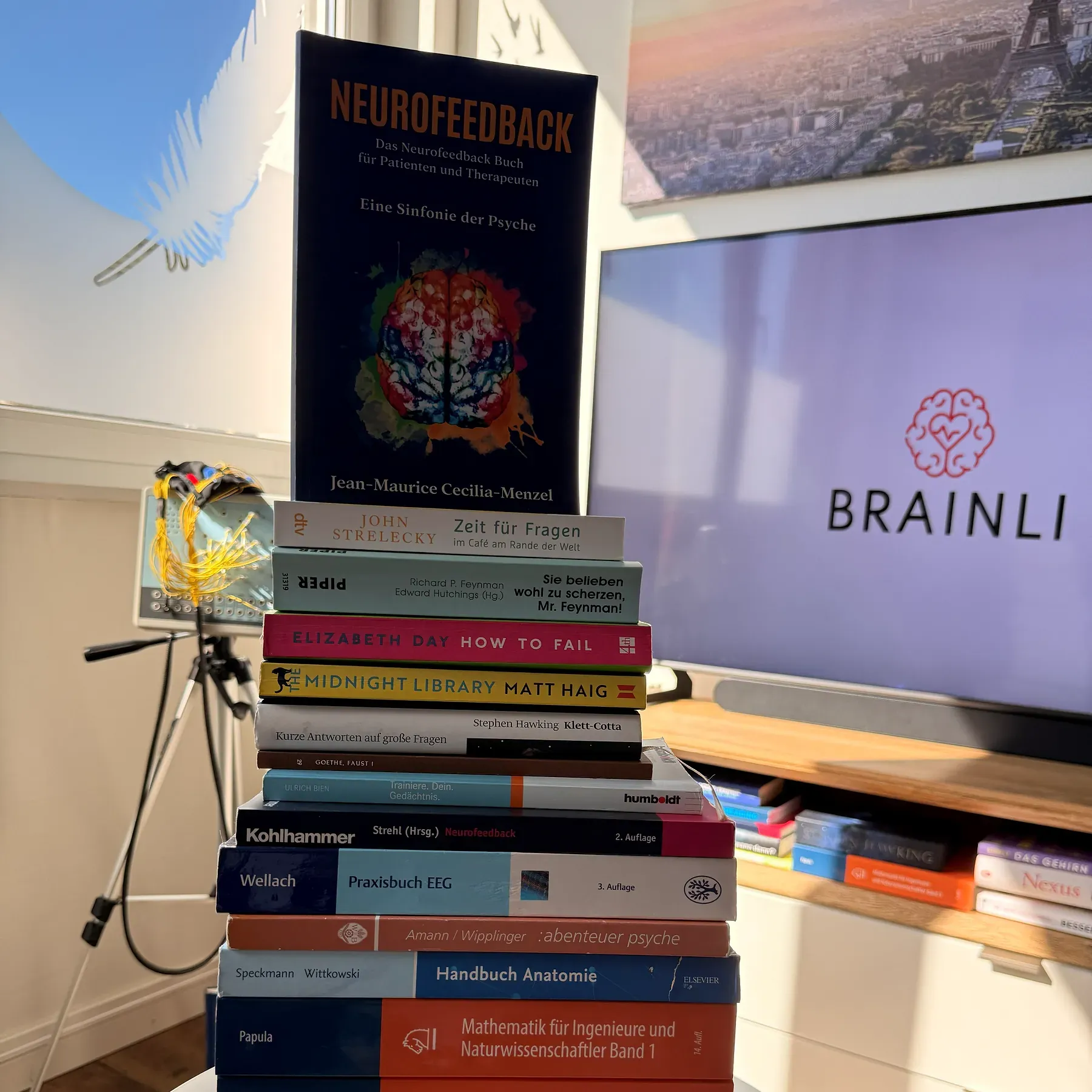 Stapel mit Fachbüchern in der Praxis, obenauf ein Buch über Neurofeedback, dahinter ein Bildschirm mit dem BRAINLI-Logo